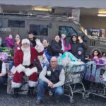 State troopers, Santa, and volunteers surrounded by carts full of toys in front of a state trooper's vehicle.