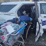A state trooper with a cart full of toys in front of his car.