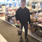 a volunteer holding up a skateboard with tables filled with toys behind him.
